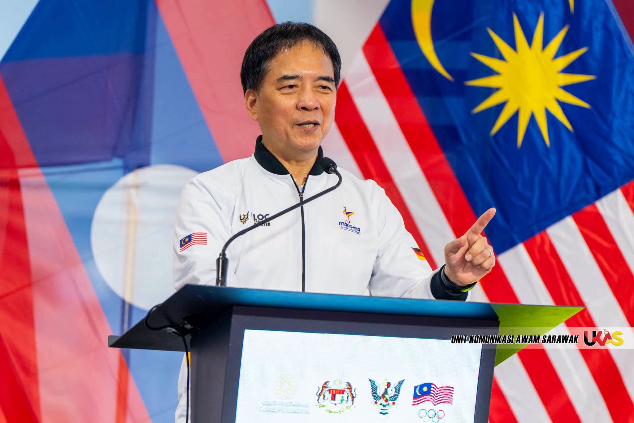 An LOC Sarawak official speaks at a lectern in front of the Sarawak flag and Jalur Gemilang.