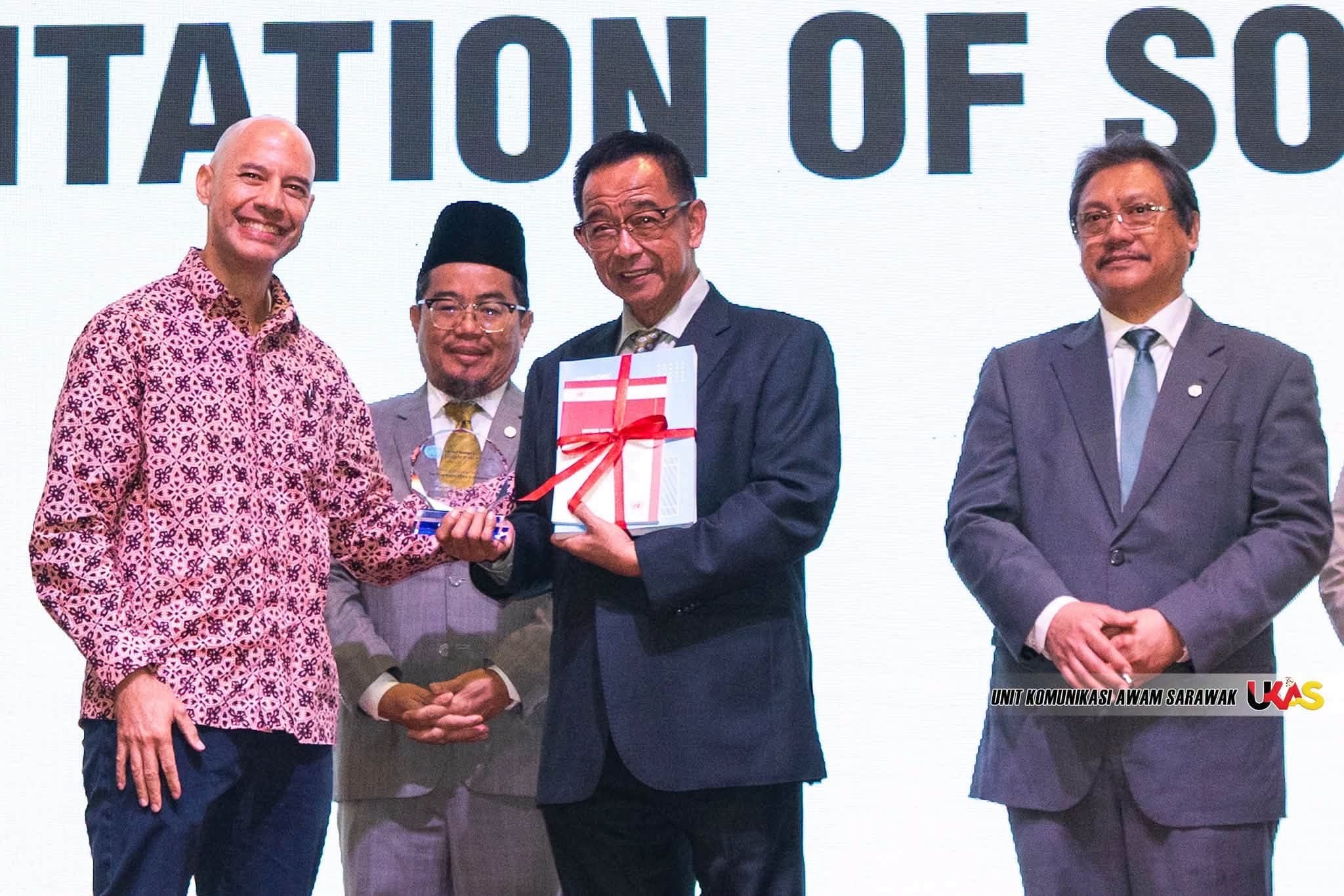 Officials on stage with a glass award and a ribboned document. The screen title is only partly in frame.